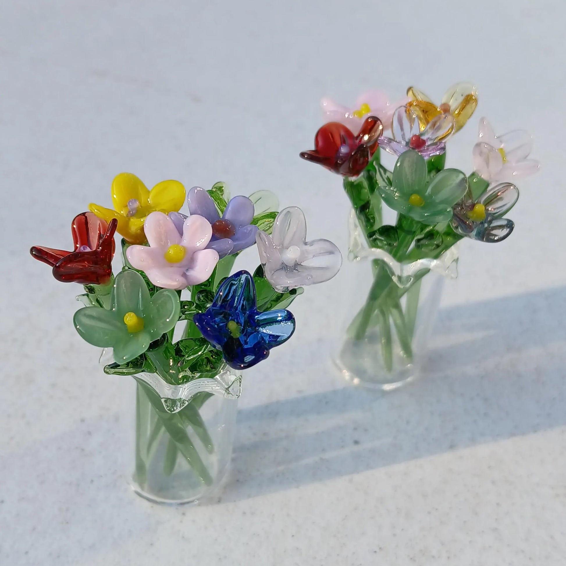 Pair of clear miniature glass vases holding vibrant handmade lampwork flowers on a light surface