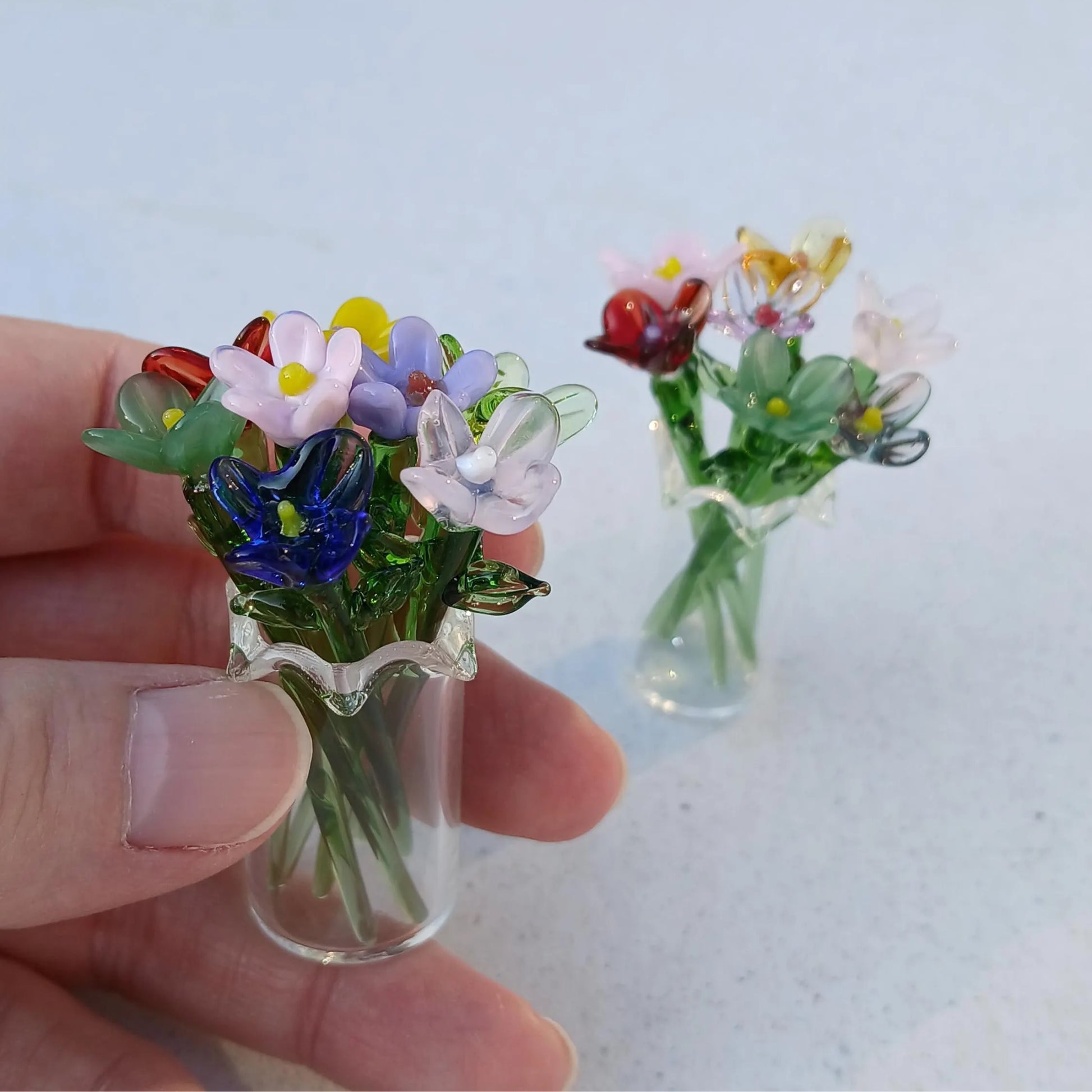 Hand holding a miniature glass vase with delicate lampwork flowers, second vase blurred in the background